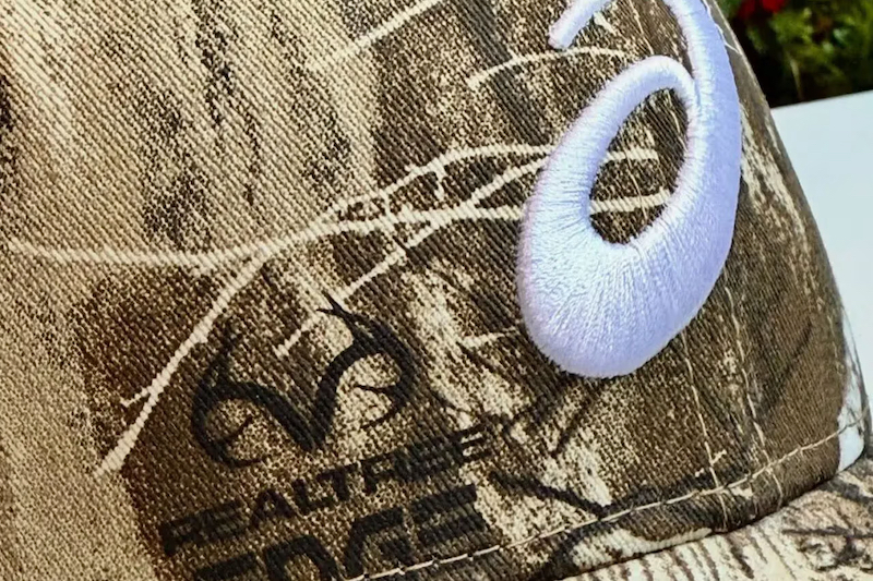 ASICSとNew Eraロゴが映えるRealtree迷彩キャップの正面デザイン。アウトドア×ストリート融合の注目アイテム。