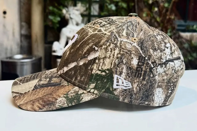 ASICSとNew Eraロゴが映えるRealtree迷彩キャップの正面デザイン。アウトドア×ストリート融合の注目アイテム。